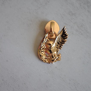 May include: A gold-colored zipper pull featuring a detailed owl design with outstretched wings. The owl is intricately crafted, showcasing feathers and facial features. The pull has a loop for attachment.