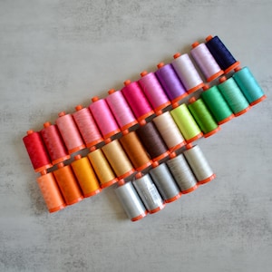 Könnte beinhalten: Draufsicht auf eine Sammlung farbiger Garnrollen, diagonal angeordnet. Die Farben reichen von leuchtendem Rot und Orange bis hin zu sanften Rosa-, Grün- und Blautönen. Jede Rolle hat einen orangefarbenen Sockel.