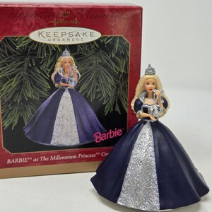 May include: A Hallmark Keepsake Barbie ornament featuring a doll dressed as The Millennium Princess. The doll has long blonde hair, a silver crown, and a dark blue gown with a glittery silver front panel. She holds a clear, spherical ornament.