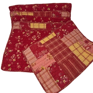 May include: A set of rectangular quilted placemats featuring a patchwork design. The fabric displays a mix of deep red floral patterns, yellow plaid squares, and striped sections. Each placemat is stitched with a grid pattern throughout.