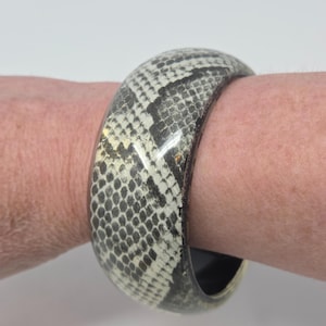 May include: A wide, chunky bangle bracelet with a snakeskin pattern in shades of black, gray, and cream. The bracelet is worn on a wrist, showcasing its glossy finish and bold design. The bracelet's diameter is approximately 3 inches.
