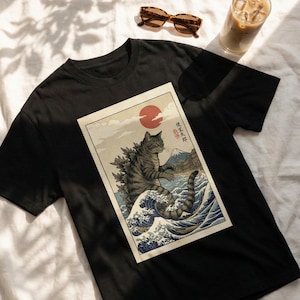 May include: Black t-shirt featuring a graphic of a cat-like monster in a Japanese woodblock print style. The print includes a red sun, waves, and a mountain. Sunglasses and a beverage are also visible.