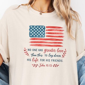 May include: Cream-colored t-shirt featuring a watercolor American flag design in red, white, and blue. The shirt includes the text "NO ONE HAS greater love than this: To lay down HIS life. FOR HIS FRIENDS. John 15:13."