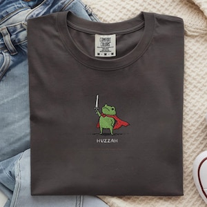 May include: Dark gray t-shirt with a cartoon frog wearing a red cape and holding a sword. The word "Huzzah" is printed below the frog. The shirt is folded on a white surface with blue jeans and white sneakers.