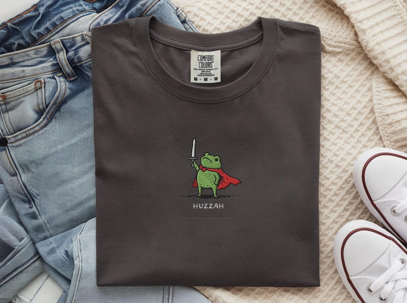 May include: Dark gray t-shirt with a cartoon frog wearing a red cape and holding a sword. The word "Huzzah" is printed below the frog. The shirt is folded on a white surface with blue jeans and white sneakers.