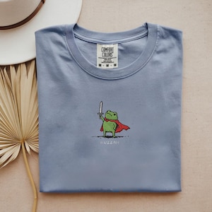 May include: A light blue t-shirt featuring a cartoon frog wearing a red cape and holding a sword. The word "Huzzah" is printed below the frog. The shirt has a crew neck and the brand "Comfort Colors" is visible on the tag.