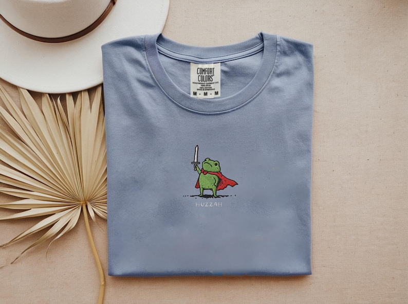 May include: A light blue t-shirt featuring a cartoon frog wearing a red cape and holding a sword. The word "Huzzah" is printed below the frog. The shirt has a crew neck and the brand "Comfort Colors" is visible on the tag.