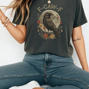 May include: Dark gray t-shirt with a raven graphic. The design features a raven perched in front of a full moon, surrounded by flowers, and the text "F-CAW-F". The shirt is a casual wear item.