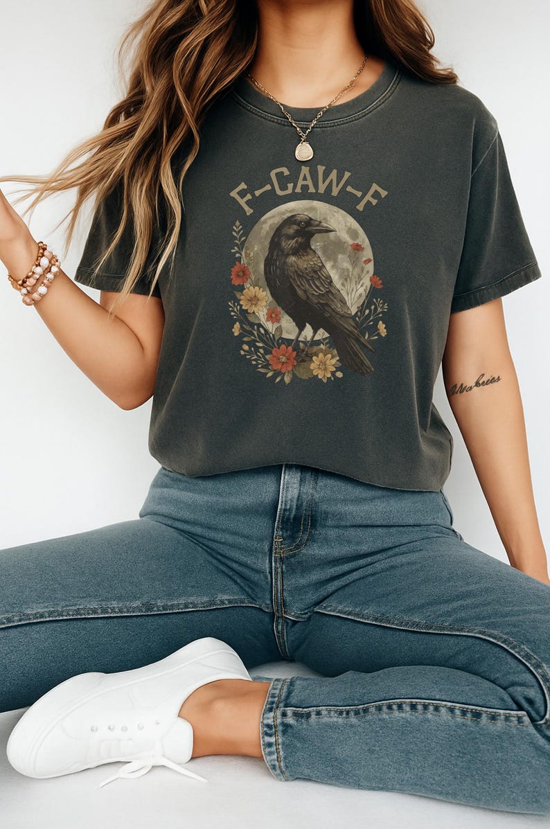 May include: Dark gray t-shirt with a raven graphic. The design features a raven perched in front of a full moon, surrounded by flowers, and the text "F-CAW-F". The shirt is a casual wear item.