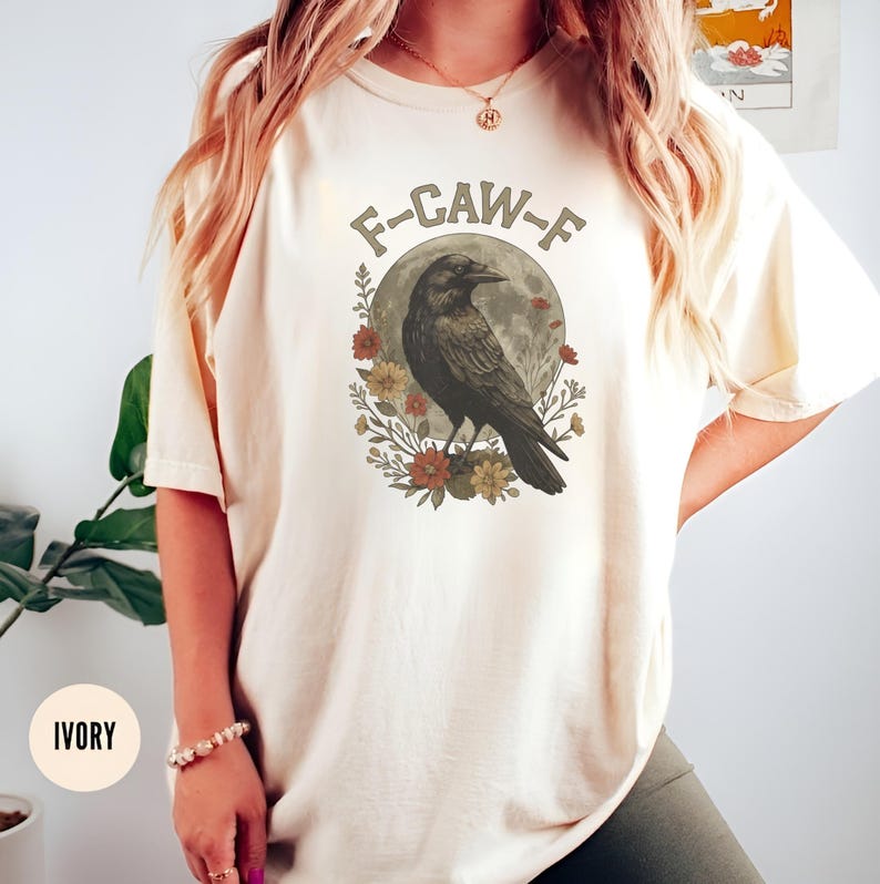 May include: Ivory-colored t-shirt featuring a graphic of a raven perched in front of a full moon, surrounded by flowers. The text "F-CAW-F" is arched above the raven. The shirt is a relaxed fit.