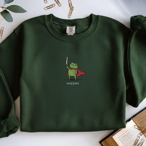 May include: Dark green sweatshirt featuring a cartoon frog wearing a red cape and holding a sword. The word "Huzzah" is printed below the frog. The sweatshirt has a crew neck and long sleeves.