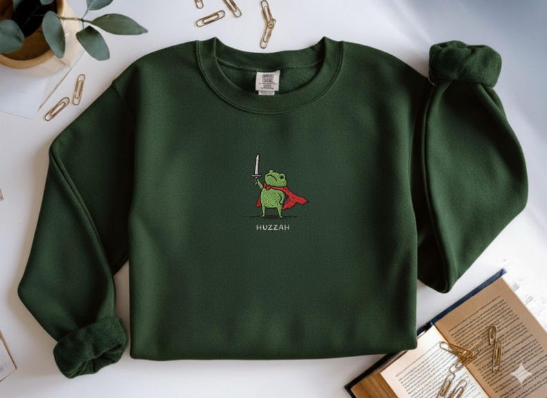 May include: Dark green sweatshirt featuring a cartoon frog wearing a red cape and holding a sword. The word "Huzzah" is printed below the frog. The sweatshirt has a crew neck and long sleeves.