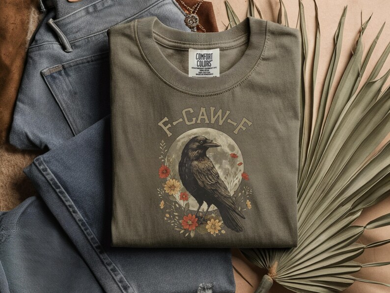 May include: Olive green t-shirt with a graphic of a black crow perched in front of a full moon, surrounded by flowers. The text "F-CAW-F" is arched above the crow. The shirt is folded on top of blue jeans.