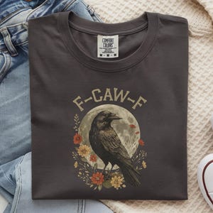 May include: Dark gray t-shirt with a round neck featuring a crow illustration. The design includes a crow in front of a full moon, surrounded by flowers, with the text "F-CAW-F" above the crow. The shirt is folded.