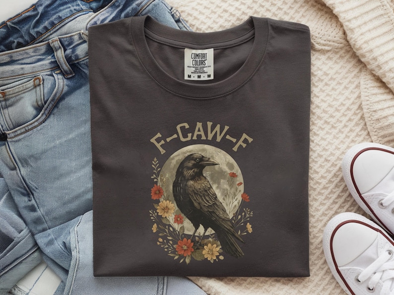May include: Dark gray t-shirt with a round neck featuring a crow illustration. The design includes a crow in front of a full moon, surrounded by flowers, with the text "F-CAW-F" above the crow. The shirt is folded.