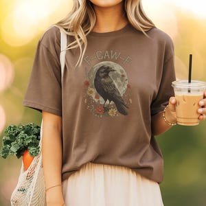 May include: Brown t-shirt featuring a black raven illustration with a full moon and floral accents. The text "F-CAW-F" is arched above the raven. The shirt is a casual style.