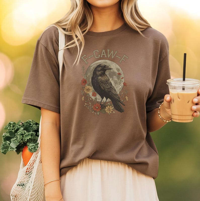 May include: Brown t-shirt featuring a black raven illustration with a full moon and floral accents. The text "F-CAW-F" is arched above the raven. The shirt is a casual style.