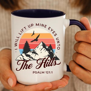 May include: White ceramic mug with a navy blue interior and handle. The mug features a mountain and sunset design with the text "I WILL LIFT UP MINE EYES UNTO THE HILLS PSALM 121:1". Two birds are flying above the mountain.