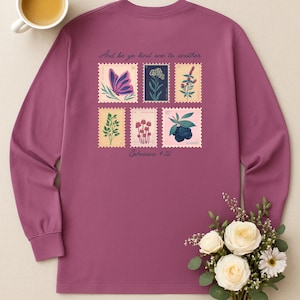 May include: A long-sleeved, mauve shirt featuring six postage stamp designs with botanical illustrations. The stamps include images of a butterfly, flowers, and leaves. The text "And be ye kind one to another" and "Ephesians 4:32" are printed on the shirt.