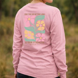 May include: A pale pink long-sleeved shirt with a floral design on the back. The design includes pink, yellow, and green flowers, along with the text "FOR THE JOY OF THE LORD IS YOUR STRENGTH" and "Nehemiah 8:10".