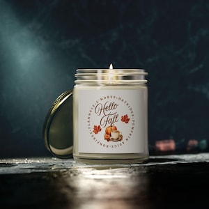 Pode incluir: Uma vela acesa em um frasco de vidro transparente com tampa prateada. O rótulo diz "Hello Fall" com uma abóbora, uma xícara de café e folhas de outono. As palavras "Flanelas, S'mores, Passeios de feno, Abóbora, Especiarias, Fogueiras" também estão no rótulo.