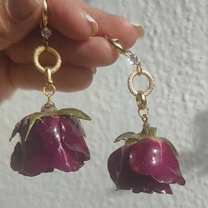 Puede incluir: Pendientes dorados con un capullo de rosa morado oscuro. Los pendientes tienen un anillo dorado texturizado y una pequeña gema transparente en la parte superior. Los capullos de rosa tienen hojas verdes en la base.