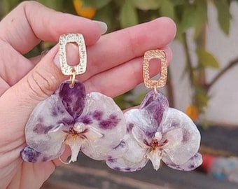 May include: A pair of handmade earrings featuring real orchid flowers preserved in clear resin. The earrings have gold-colored rectangular top pieces. The orchid petals are white with purple spots and accents.