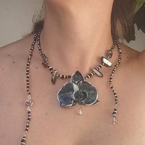 May include: A handcrafted necklace featuring a large, dark gray orchid pendant with silver accents. The necklace includes strands of small, dark beads and larger, irregularly shaped stones. A small silver charm hangs from the orchid.