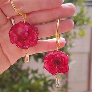 Puede incluir: Pendientes de aro dorados con colgantes de rosas rojas vibrantes. Cada pendiente tiene un dije dorado en forma de lágrima colgando debajo de la rosa. Los pendientes se muestran sobre un fondo verde borroso.