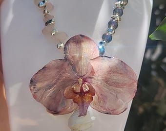 May include: A necklace featuring a large, dried pink orchid pendant with a white seashell and a beaded chain. The chain alternates between light pink stone beads and silver-toned metal beads, and blue crystal beads.