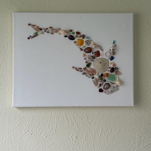 Puede incluir: Una obra de arte en lienzo blanco que presenta un diseño de pez hecho de varias conchas marinas, vidrio de mar y pequeñas piedras. La obra de arte está montada sobre un lienzo blanco y cuelga contra una pared texturizada de color claro.
