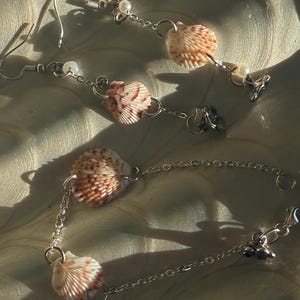 Puede incluir: Pendientes y pulsera de conchas marinas. La joyería presenta pequeñas conchas marinas naturales con marcas rojizas, ensartadas en delicadas cadenas de plata. Pequeñas perlas blancas y dijes decorativos añaden detalles.