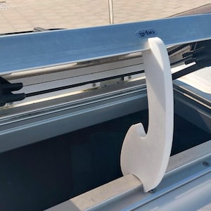 May include: A white, curved support bracket is attached to a silver metal frame, possibly for a boat or vehicle. The bracket is positioned near a dark opening, with blue accents visible. The word "gebo" is printed on the frame.