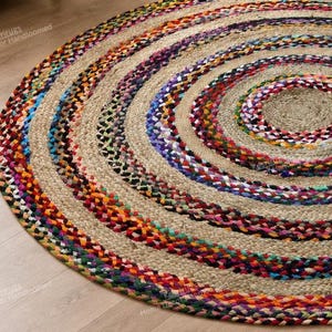 May include: A round braided rug featuring concentric circles of natural tan jute fibre alternating with vibrant multi-coloured fabric strips in shades of red, blue, yellow, green, and purple. The rug has a textured, woven appearance throughout.