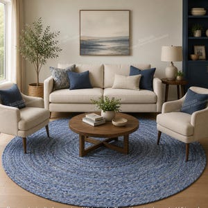 May include: A large, round, braided area rug in shades of blue and grey, featuring a thick, textured weave. The rug is positioned on a light-coloured floor beneath a wooden coffee table, two beige armchairs, and a matching beige sofa with blue pillows.