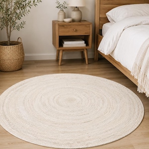 Può includere: Un tappeto rotondo color bianco sporco con un motivo intrecciato a cerchi concentrici. Il tappeto è realizzato in materiale intrecciato e testurizzato e poggia su un pavimento in legno chiaro, parzialmente sotto una struttura letto in legno.