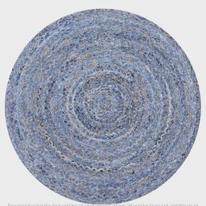 Op de afbeelding: Een rond vloerkleed met een gevlochten constructie gemaakt van diverse tinten blauwe en lichtgrijze stoffen stroken. Het cirkelvormige ontwerp toont een getextureerd, geweven patroon met concentrische ringen vanuit het midden naar de rand.