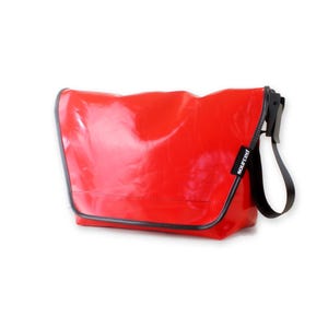 Puede incluir: Un bolso de mensajero rojo brillante con una correa negra y un ribete negro. El bolso tiene una etiqueta negra "Scout" en la parte delantera.