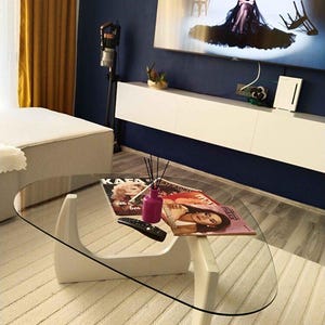 May include: A contemporary living room scene with a white coffee table. The table has a clear glass top and a distinctive curved base. Magazines and a remote control are on the table. A TV displays an image, and a diffuser is present. The room has a rug.