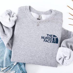 Könnte beinhalten: Ein hellgraues Sweatshirt mit dem navyblauen Schriftzug "THE NURSE FACE" und einem medizinischen Symbol. Die Ärmel haben ein weißes Futter. Das Bild zeigt auch eine hellblaue zerrissene Jeans und eine silberne Halskette. Das Sweatshirt ist wahrscheinlich aus weichem Material.