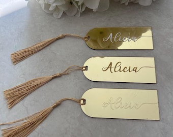 May include: Three gold-colored name tags with the name "Alicia" engraved in a cursive font. Each tag has a matching beige tassel attached with a string. White flowers are in the background.