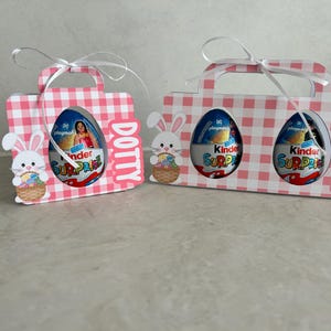 May include: Two pink and white gingham Easter gift boxes, one with the name "Dotty" and the other with "Ava". Each box has a handle and a cutout showing a Kinder Surprise egg. Bunny and basket cutouts are attached.