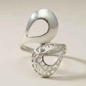 May include: A silver ring featuring two asymmetrical teardrop-shaped loops. The upper loop has a smooth, polished surface, while the lower loop displays an intricate, openwork lattice pattern that resembles a delicate, interconnected web structure.