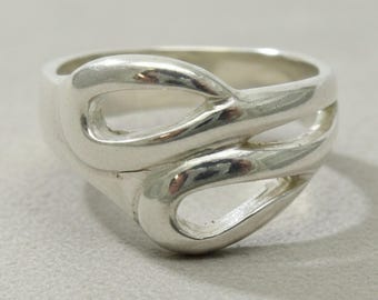 Puede incluir: Un anillo de plata pulida con una banda fluida y continua que se entrelaza para crear dos aberturas ovaladas asimétricas. La superficie metálica es lisa y reflectante, mostrando un diseño único e interconectado que rodea el dedo.