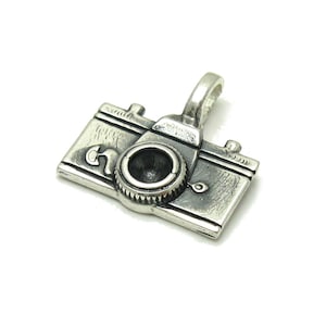 May include: A silver charm shaped like a vintage camera. The charm features a detailed design with a lens and a small loop at the top for attaching to a bracelet or necklace.