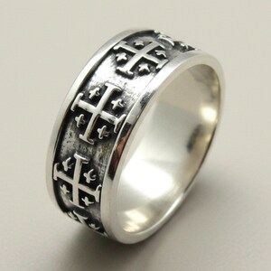 Könnte beinhalten: Ein silberner Ring mit einem Band, das ein sich wiederholendes, geprägtes Jerusalemkreuz-Muster aufweist. Das Metall hat polierte Kanten und einen abgedunkelten, oxidierten Hintergrund, um die erhabenen Kreuzdetails rundherum hervorzuheben.