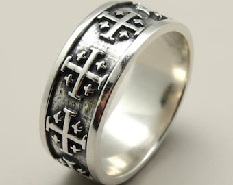 May include: A silver ring featuring a band with a repeating embossed Jerusalem cross pattern. The metal has a polished finish on the edges and a darkened, oxidized recessed background to highlight the raised cross details around the entire circumference.
