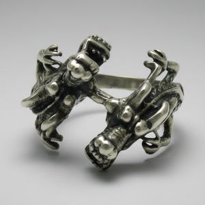 May include: A silver-toned metal ring featuring two detailed dragon heads facing each other. The band is open in the front, with the dragon bodies forming the sides of the ring. The metal has a polished finish with dark recessed areas to highlight the scales.
