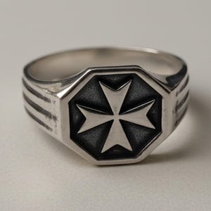 Könnte beinhalten: Ein Ring aus silberfarbenem Metall mit einer achteckigen Vorderseite, die ein vertieftes, dunkel getöntes Malteserkreuz-Emblem zeigt. Das Band weist auf jeder Seite drei parallele horizontale Rillen auf, die der polierten Oberfläche Struktur verleihen.