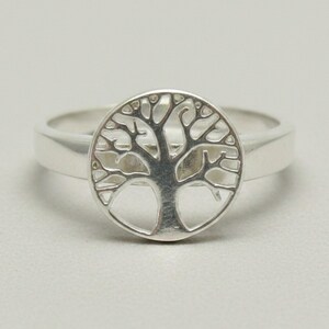 May include: A polished silver ring featuring a circular centerpiece with a cut-out design of a tree with spreading branches and a solid trunk. The band is smooth and tapers slightly toward the back, showcasing a metallic finish throughout.
